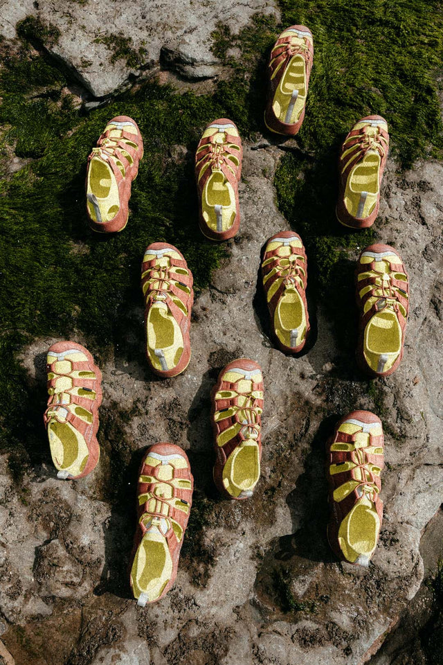 Serie di sandali Keen Hyperport H2 patrol in colore arancione e giallo disposti su una superficie rocciosa con muschio verde, visti dall’alto. Le scarpe sono allineate in modo regolare, creando un pattern visivo tra texture naturale e design outdoor.