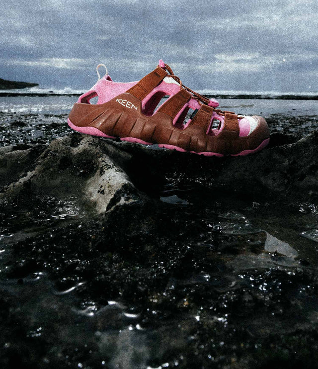 Sandalo Keen Hyperport H2 in colore rosso e rosa appoggiato su una roccia umida in riva al mare, con il cielo nuvoloso sullo sfondo. L’immagine mette in risalto il design robusto e la suola adatta a terreni bagnati e scivolosi.
