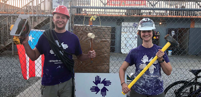 Due volontari sorridenti con attrezzi da lavoro e magliette con il logo di "All Hands and Hearts Smart Response" in un progetto di ricostruzione a Porto Rico.