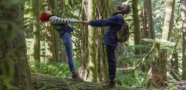 Coppia felice in escursione nella foresta, che si tiene per mano tra gli alberi secolari – indossano scarponcini da trekking KEEN e abbigliamento outdoor.