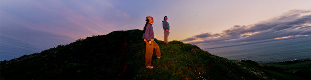 Due persone con scarpe KEEN Targhee IV camminano su una collina affacciata sul mare al tramonto.