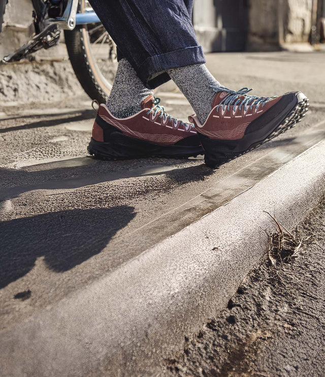 Dettaglio di scarpa KEEN Zionic Collab. Pilgrim Surf Supply su sabbia, accanto a una tavola da surf. Il modello combina design tecnico e stile outdoor, ideale per esplorare anche ambienti costieri. Suola resistente e tomaia robusta garantiscono aderenza e comfort, perfetti per chi ama muoversi tra natura e avventura urbana.