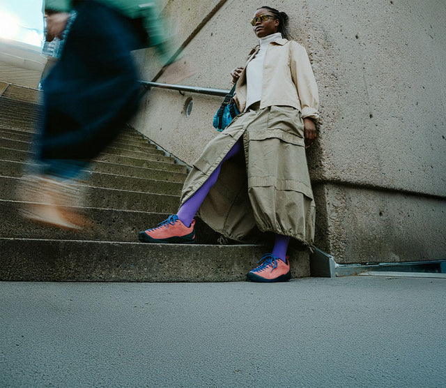 Donna con scarpe KEEN Jasper da donna in tonalità corallo e blu, fotografata su una scala cittadina in look streetwear outdoor. Scarpe versatili e comode per uso urbano e avventure quotidiane.