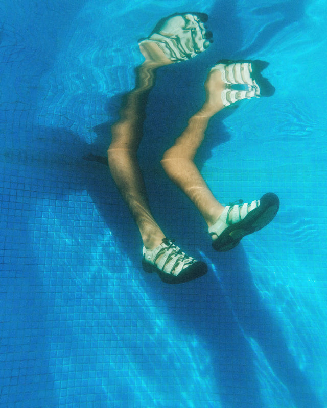 Una persona con indosso i sandali Keen Newport Retro in canapa è immersa in piscina. Le gambe e le calzature sono riflesse e distorte dall’acqua limpida, creando un effetto visivo onirico e dinamico. L’immagine enfatizza la natura anfibia del sandalo e lo stile libero della collezione Gramicci x KEEN.