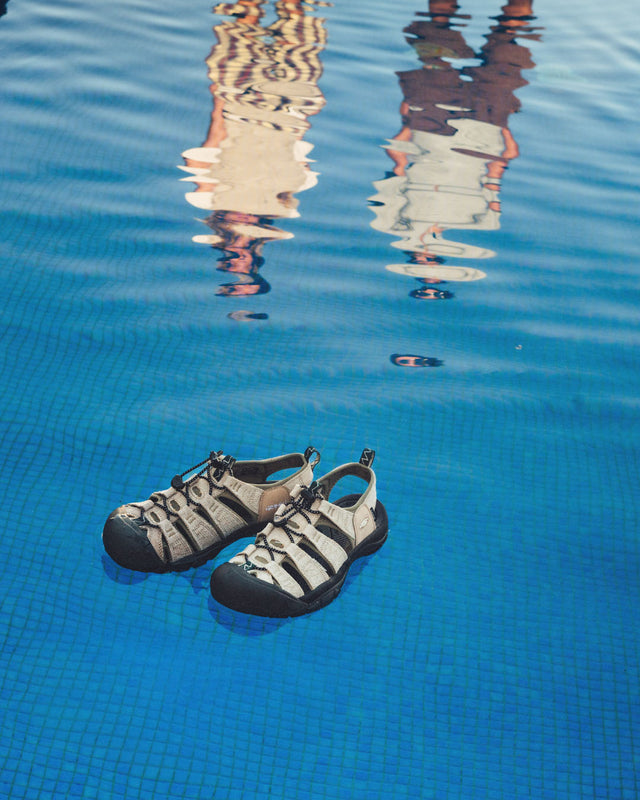 Un paio di sandali Keen Newport Retro in canapa galleggiano perfettamente sull'acqua di una piscina, mentre sullo sfondo si riflettono le sagome distorte di due persone. L’immagine trasmette leggerezza, libertà e sostenibilità, in linea con lo spirito outdoor della collaborazione tra KEEN e Gramicci.