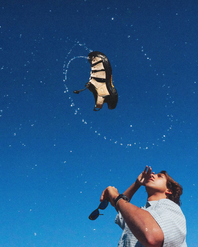Un giovane uomo guarda in alto mentre un sandalo Keen Newport Retro in edizione limitata, realizzato in canapa, fluttua nel cielo blu con un effetto dinamico d’acqua. La scena celebra l’unione tra avventura e sostenibilità, simbolo della collaborazione tra KEEN e Gramicci.