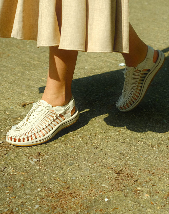 Dettaglio delle gambe di una donna che cammina su un sentiero asfaltato indossando sandali Keen Uneek in colore beige, abbinati a una gonna chiara, in una giornata di sole.