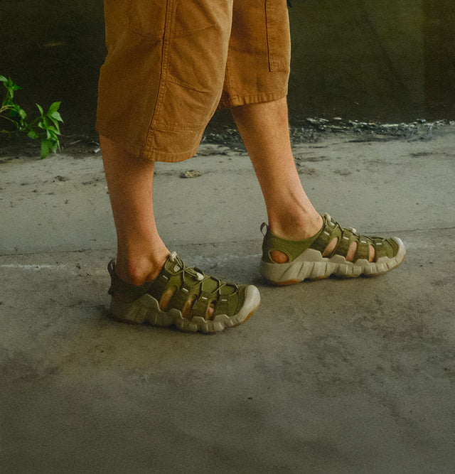 Dettaglio di una persona che cammina indossando sandali Keen Hyperport H2 da uomo in colore verde oliva, su una superficie in cemento in un ambiente urbano o semi-naturale.