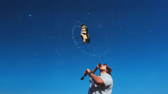 Un giovane uomo guarda in alto mentre un sandalo Keen Newport Retro in edizione limitata, realizzato in canapa, fluttua nel cielo blu con un effetto dinamico d’acqua. La scena celebra l’unione tra avventura e sostenibilità, simbolo della collaborazione tra KEEN e Gramicci.