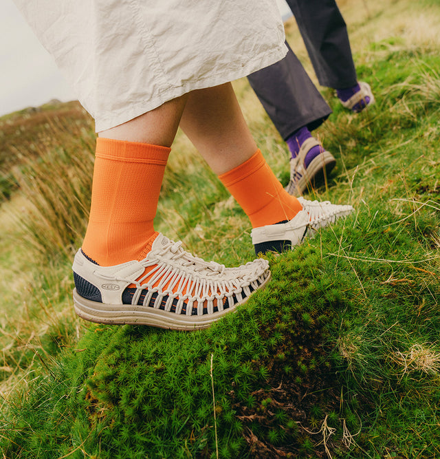 Due persone camminano su un prato verde indossando sandali KEEN UNEEK, perfetti per attività outdoor. Il design intrecciato si adatta al piede offrendo comfort e flessibilità. La combinazione di calzini colorati e natura esalta lo stile lifestyle del modello, pensato per chi ama esplorare con leggerezza e originalità.