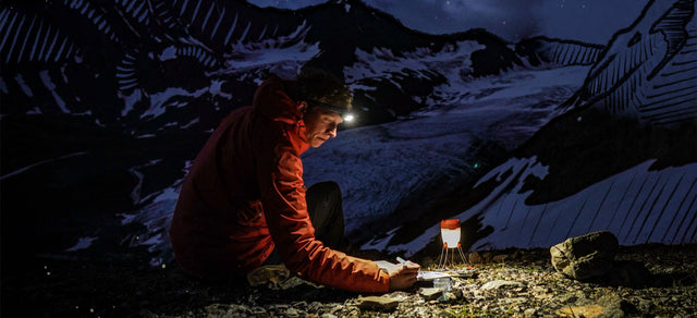 Persona con giacca rossa e lampada frontale accesa scrive vicino a una lanterna durante un'escursione notturna in montagna.
