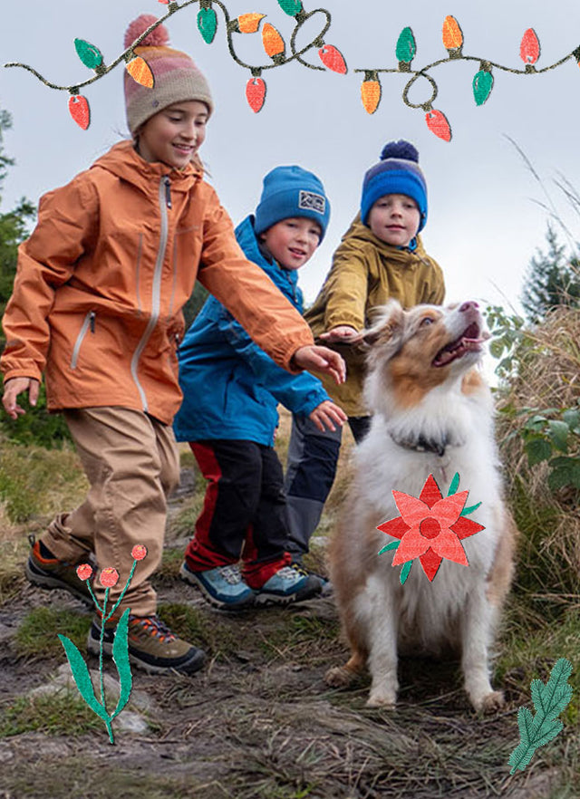 Gruppo di bambini che camminano all’aperto con scarponcini KEEN, insieme a un cane, su sfondo decorazioni natalizie illustrate.