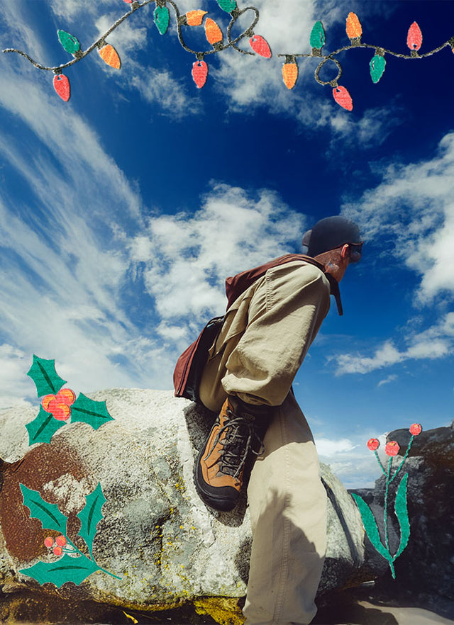 Uomo seduto su una roccia in montagna con scarponcini KEEN marroni, cielo blu e decorazioni natalizie illustrate.